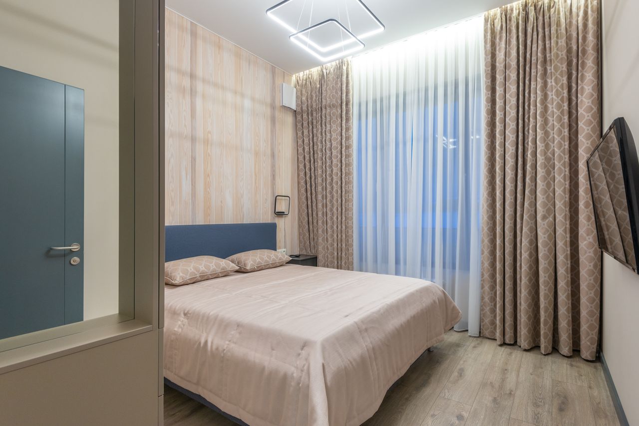 services-05 Comfortable bed with cushions and TV hanging on wall in cozy bedroom decorated with curtains