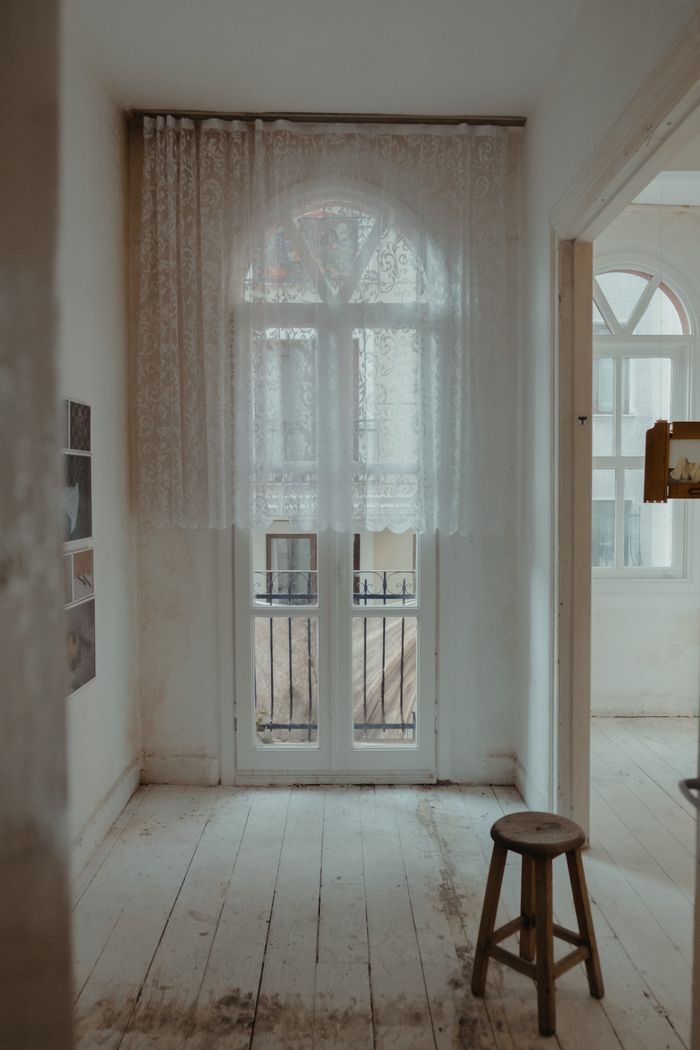 portfolio-02 A tranquil indoor scene with lace curtains and a wooden stool by bright windows.